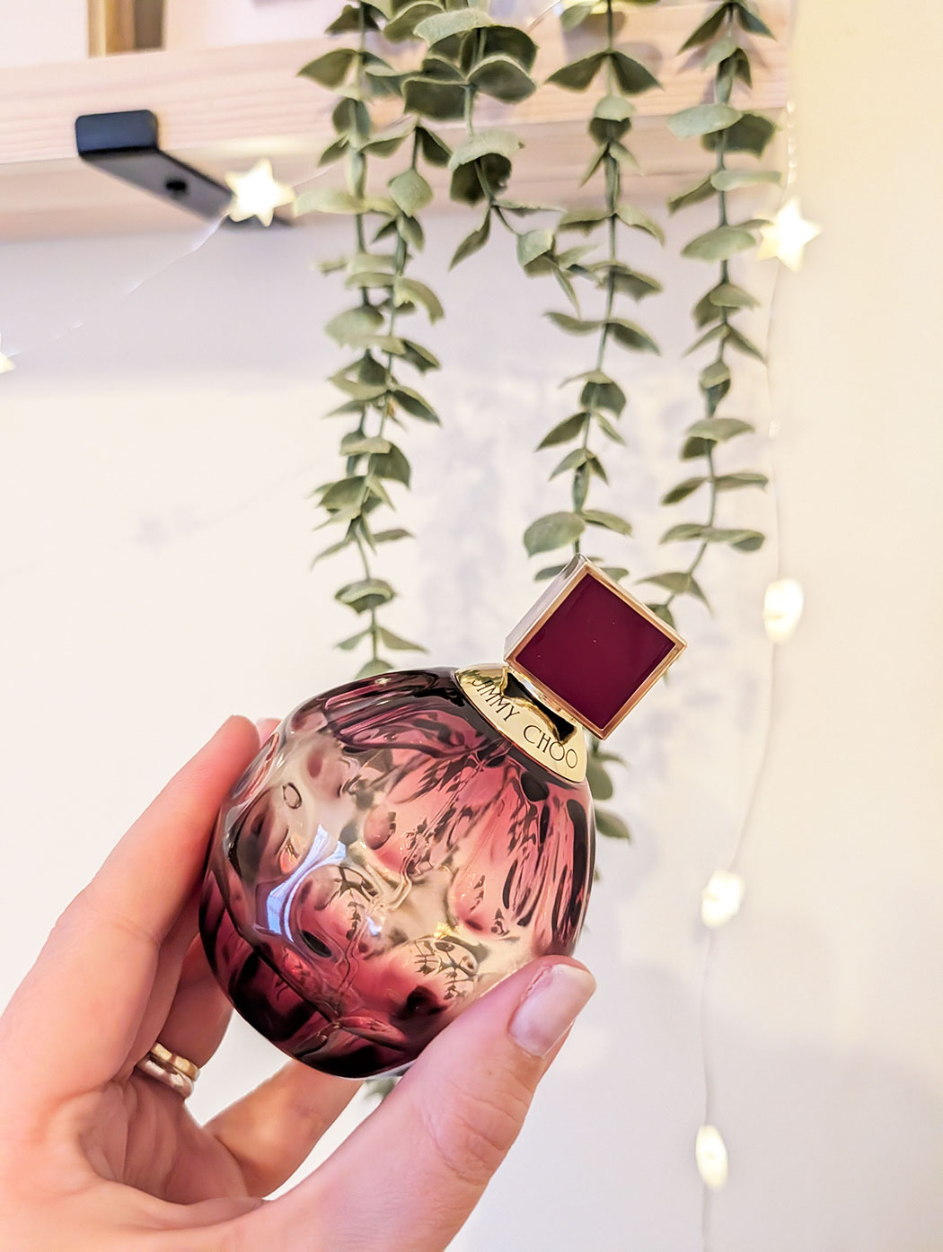 Jimmy Choo fragrance being held by a women's hand against a string plant and twinkle lights
