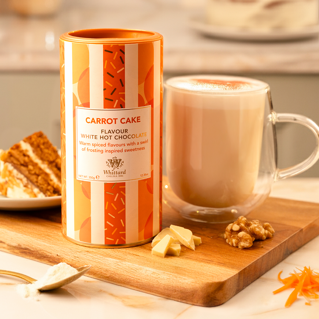 Carrot cake hot chocolate in orange and white sriped cannister next to a cup of frothy hot chocolate