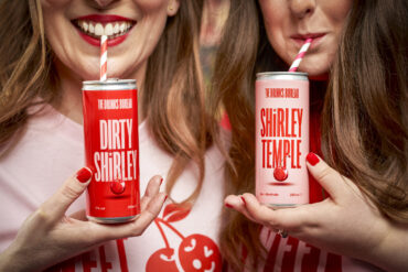 Two women drinking dirty shirley drinks from pink and red cans via a straw