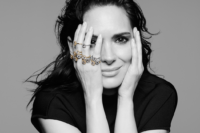 Black and white image of actress Winona Ryder with jewellery on each of her fingers and both hands pressed against her face.